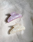 Two bunny-shaped hair clips, one purple and one white, on a light fabric background.