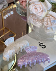Two hair clips, one beige and one purple glittery, on a glass surface with flowers in the background.